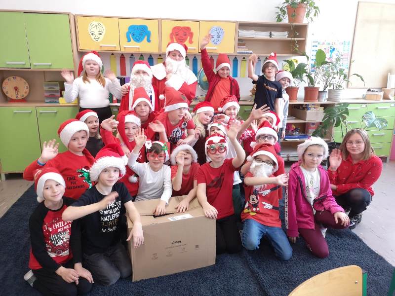 Uczniowie klasy 2b w mikołajkowych czapkach wesoło pozują na dywanie do zdjęcia z Mikołajem oraz reniferem, którzy przynieśli im skrzynię z prezentami.
