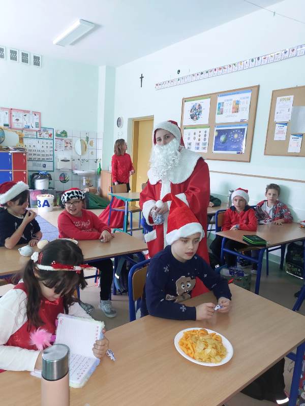 Mikołaj oraz renifer są w klasopracowni klasy 3a - Uczniowie siedzą przy swoich ławkach, Mikołaj oraz renifer przechadzają się między ławkami częstując ich słodkościami.