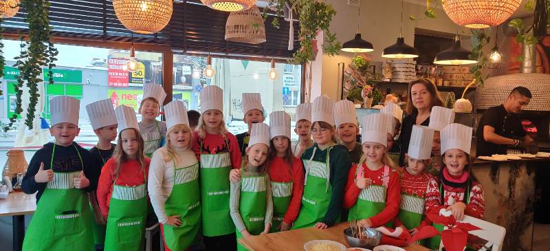 Dzieci stoją w grupie, ubrane w zielone fartuszki i czapeczki kucharskie, z tyłu wnętrze pizzerii.