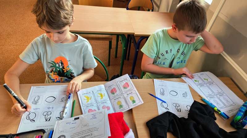 Dwaj uczniowie siedzą w ławce, kolorują owoce i podpisują je w języku francuskim.