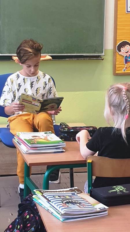 Uczeń w żółtych spodniach siedzi na niebieskim krześle, czyta książkę.