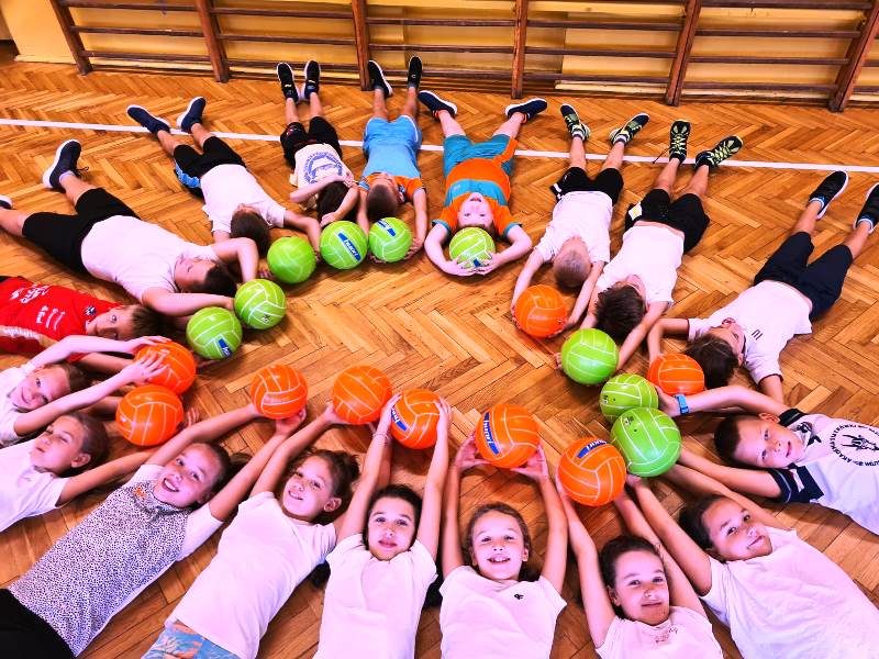 Klasa 3b, dzieci leżą kołem na podłodze w sali gimnastycznej, nad głowami trzymają kolorowe piłki.